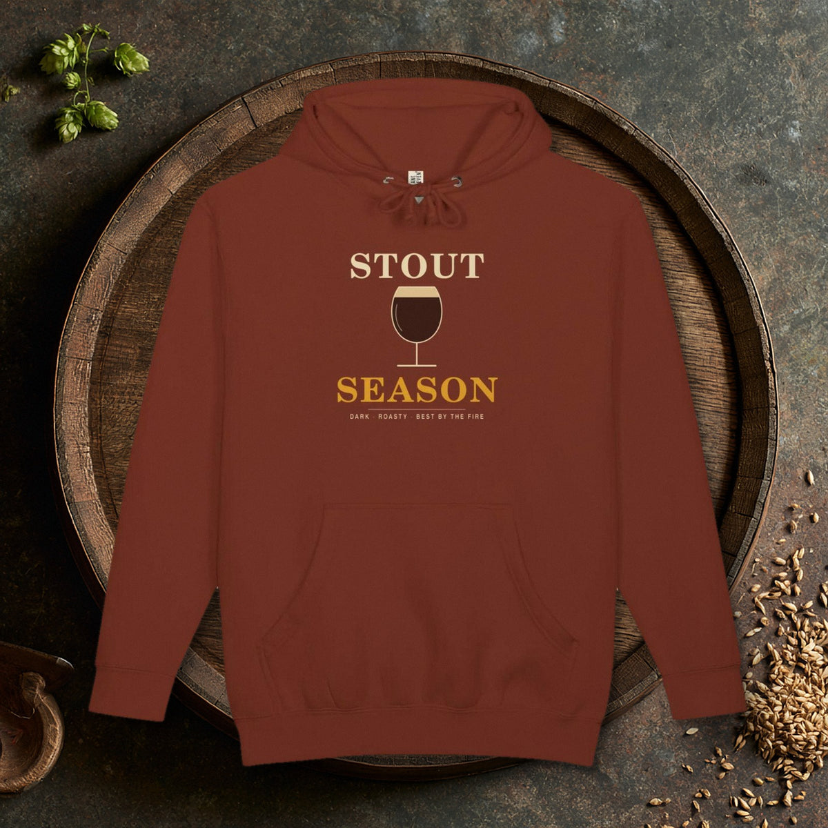 Stout Season Heavyweight Hoodie in Paprika laid flat on an oak barrel top with hops and malt