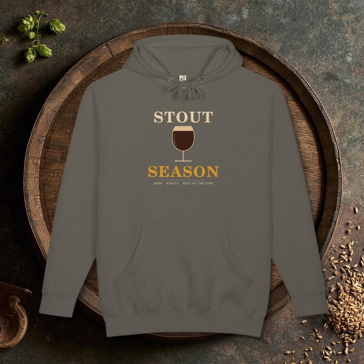 Stout Season Heavyweight Hoodie in Storm laid flat on an oak barrel top with hops and malt