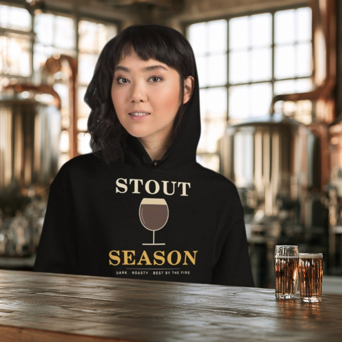 Woman in the black Stout Season hoodie at a brewery bar with two pints and fermenters behind