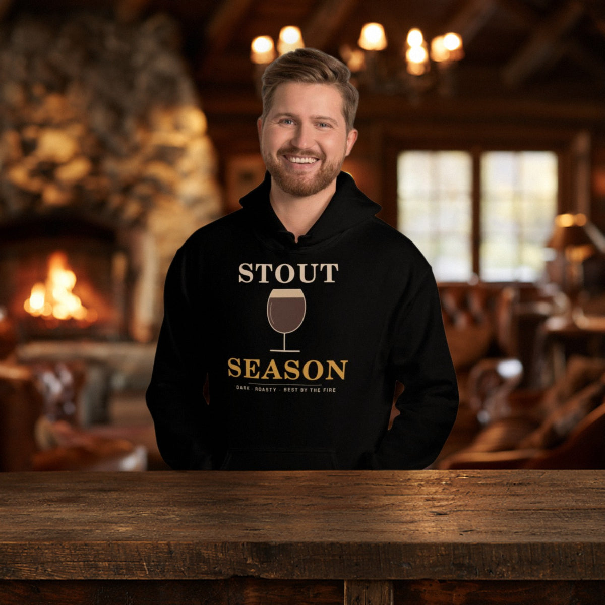 Smiling man in the black Stout Season hoodie at a rustic lodge with a stone fireplace burning behind him
