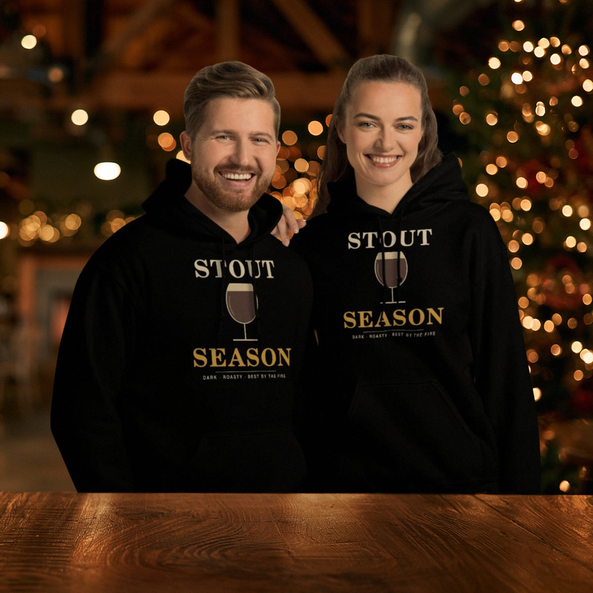 Couple in matching black Stout Season hoodies at a festive taproom with a lit Christmas tree